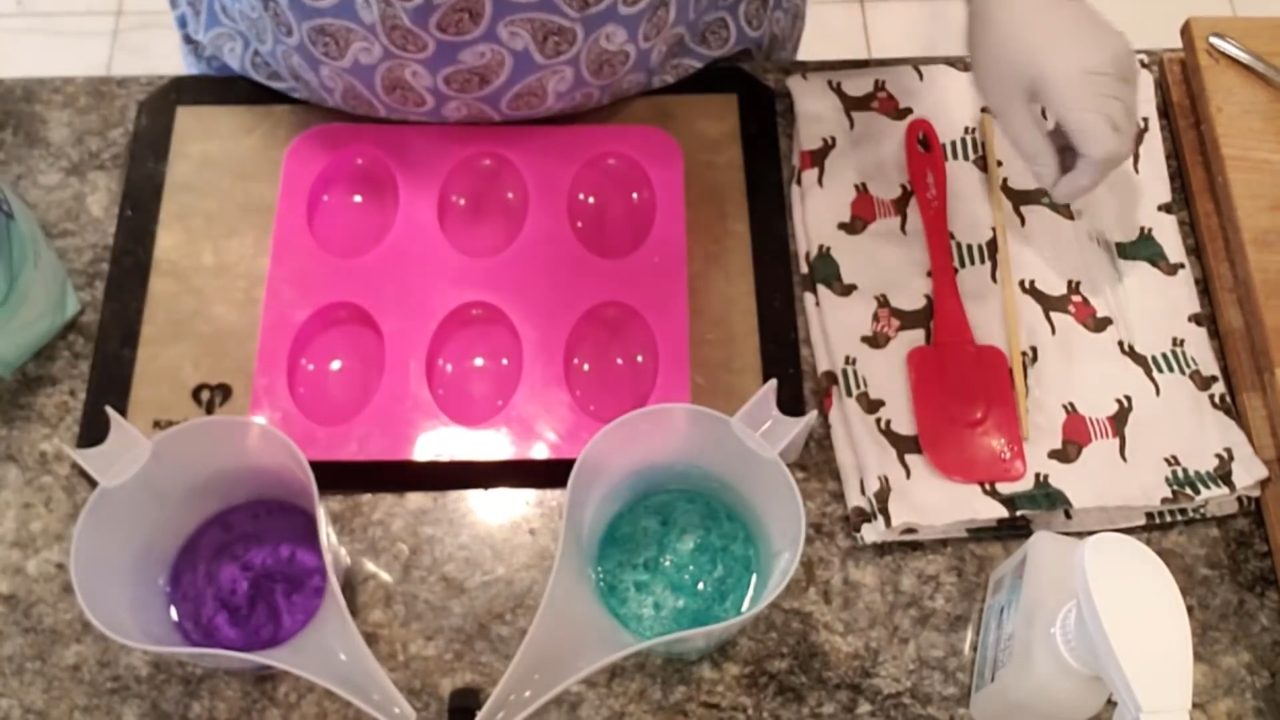 Purple and green melted clear soap in pour cups next to an oval mold